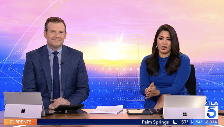 Two news anchors presenting on a television broadcast with a cityscape in the background.
